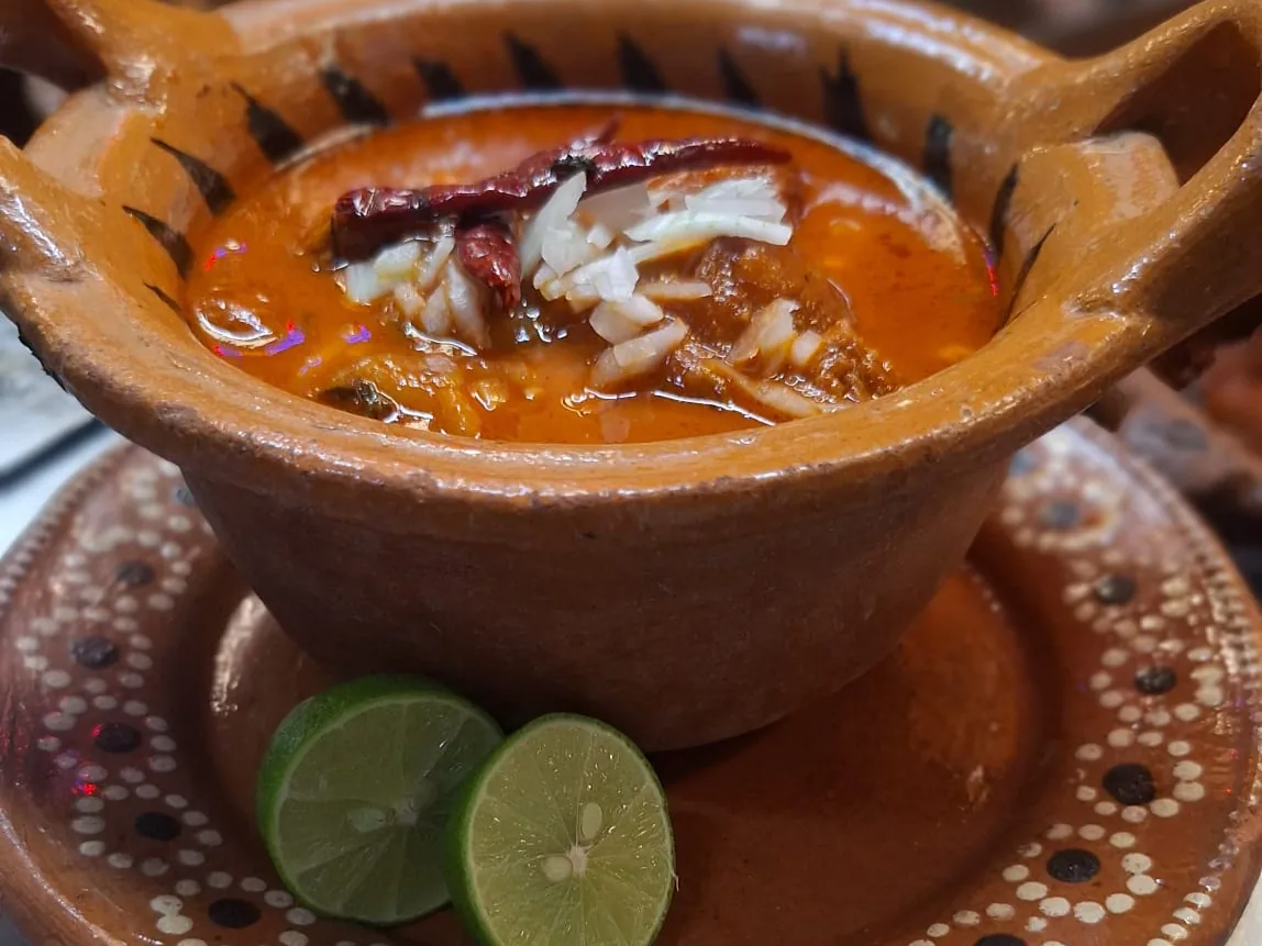 Cazuela de barro con menudo rojo, cebolla, chile de árbol y limones